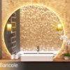 Circle-shaped warm backlit mirror with two bancole lamps on a gold mosaic wall in a washroom.