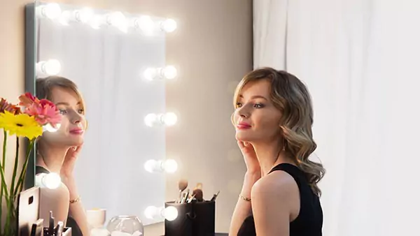 Preview of woman gazes at her reflection in a vanity mirror with warm light bulbs, captivating her own beauty and radiance.