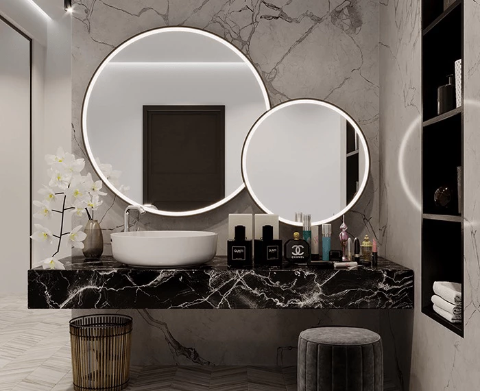 A bathroom with marble walls and two different-size round-lighted mirrors above a single basin.